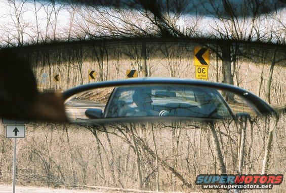 Reflection of car interior and driver in side mirror, with trees and road signs visible