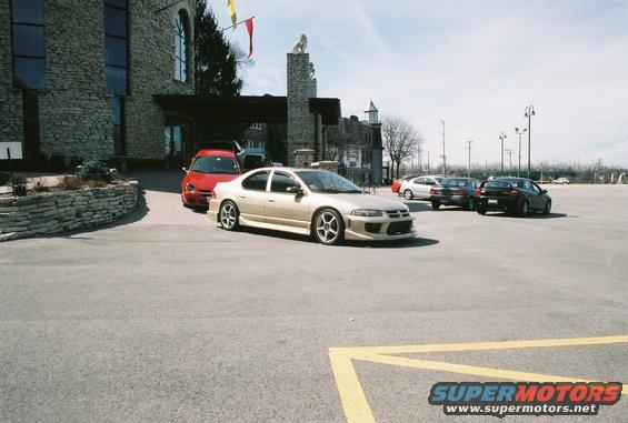 Gold sedan with aftermarket body kit and wheels parked in a lot near a stone building. Red car and other vehicles are also visible
