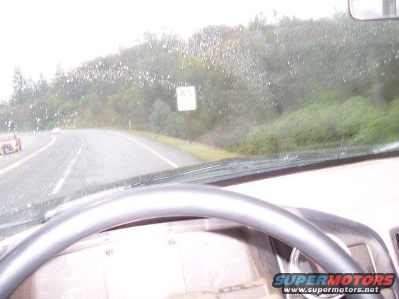 View from inside a vehicle driving on a wet road with trees and a speed limit sign visible through the windshield