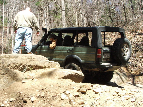 Green SUV navigating rocky terrain with a person standing on a boulder