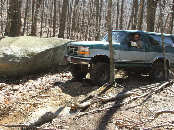 Green and silver 1996 Ford Bronco driving over rocks and logs in a wooded area