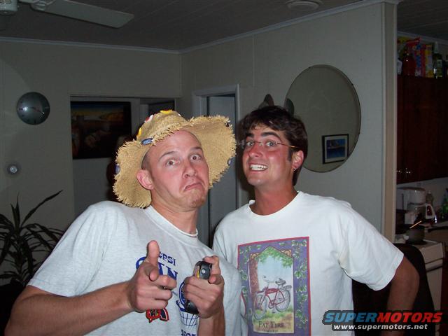 Man in straw hat points finger gun, man in glasses smiles, indoor scene with kitchen counter and cabinets