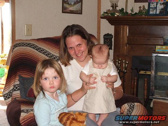 Woman smiles holding a baby while a young girl sits beside her on a couch