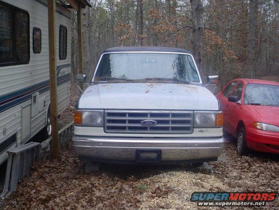 White Ford Bronco with a dark roof, front view, showing a chrome grille with a Ford emblem, parked next to an RV and a red sedan