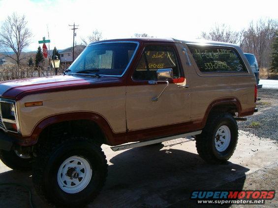 1985 Ford Bronco, tan and maroon two-tone paint, lifted with large tires. "For Sale" and phone number written on windows
