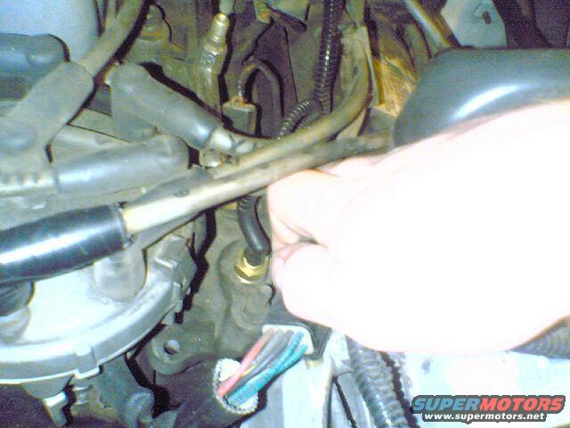 Close-up of engine bay with hand reaching for sensor. Wires and hoses visible