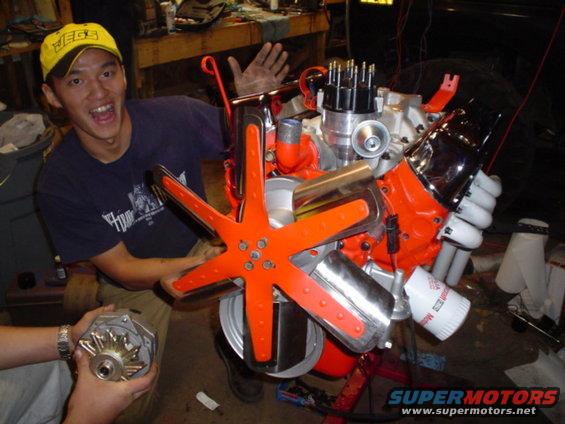 Orange V8 engine with chrome accents and a large orange fan, held by a person wearing a yellow cap