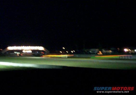 Blurry car lights streak across a dark track at night, with a lit structure in the background