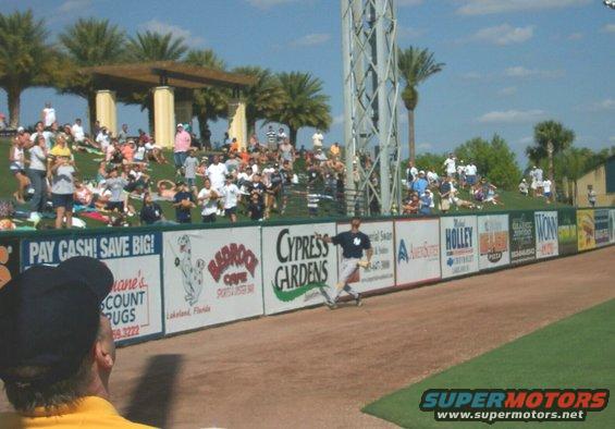 Spectators watch a baseball game from grassy berms behind a wall with advertisements for Cypress Gardens, Holley, and others