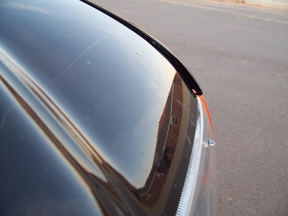 Black car hood reflecting a brick building, with an orange tow strap near the headlight
