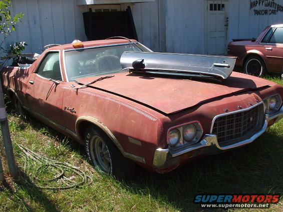 Red 1972 Ford Ranchero with a chrome trim piece on the hood, parked in grass next to a building
