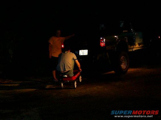 Person in red wagon pulled by another person next to a dark-colored truck at night
