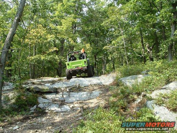 Green off-road vehicle with two seats, orange flag, and aftermarket bumper driving on a rocky trail