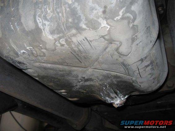Underside of a vehicle's fuel tank with dents and scratches, showing exhaust pipes below