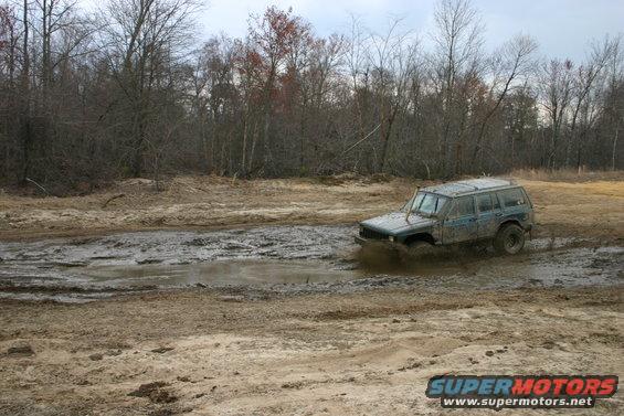 Muddy blue Jeep Cherokee XJ driving through a muddy puddle off-road