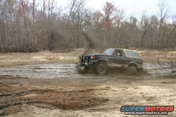 1989 Ford Bronco driving through mud with trees in background