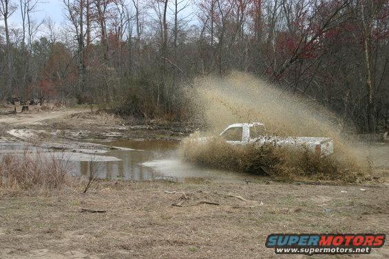 White pickup truck driving through a muddy puddle, kicking up a lot of water and dirt
