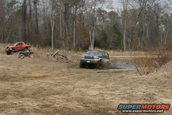 1989 Ford Bronco driving through a muddy puddle off-road, with an orange pickup truck and ATV visible in the background