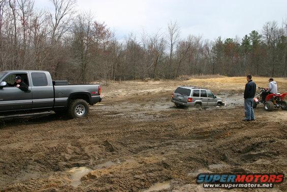 Gray pickup truck and Jeep stuck in mud, with two people watching and an ATV nearby
