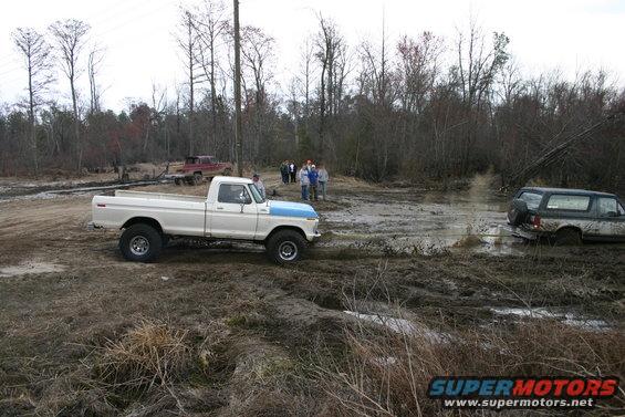 White and blue Ford pickup truck on dirt, with a Bronco stuck in mud in the background