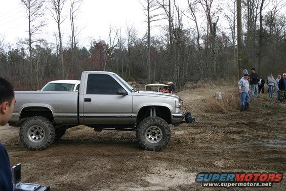 Lifted silver pickup truck with large tires parked in mud; another vehicle and people in background