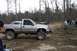 1989 Ford Bronco - Silver pickup truck with lifted suspension and large off-road tires sits in a muddy area with trees and people around.