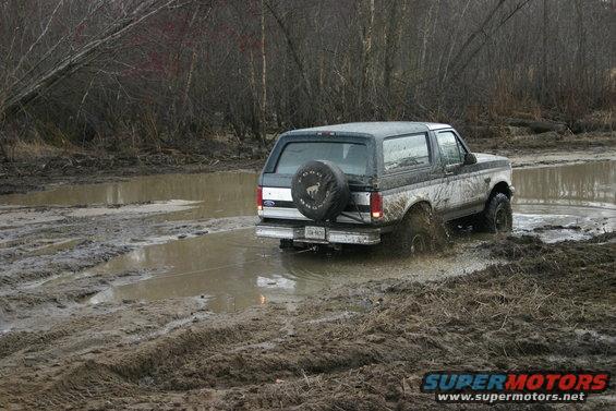 Ford Bronco driving through a muddy trail with trees in the background