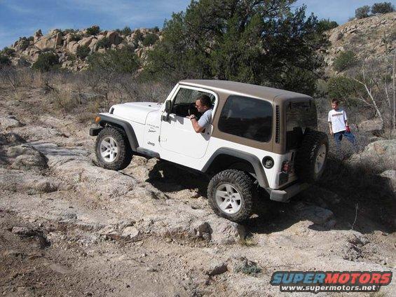 White Jeep TJ driving over rocks with a man inside and a kid standing nearby