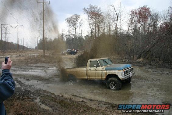 Ford pickup truck with blue hood driving through mud, kicking up spray. Another vehicle is visible in the background