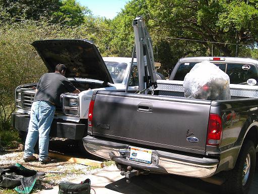 Man leans into engine bay of a white Ford F-Series truck, possibly 1980-1986. A dark gray Ford F-Series truck is parked next to it