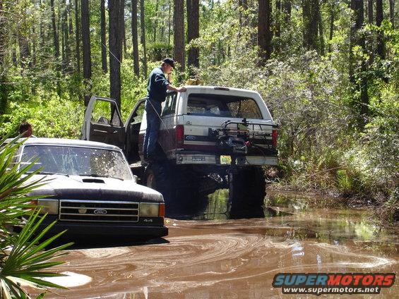 Brown and white Ford Bronco stuck in mud with a person standing on the rear bumper
