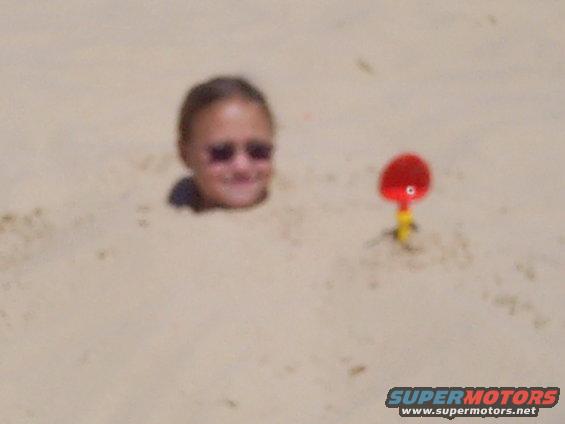 Child with sunglasses buried in sand next to a red toy