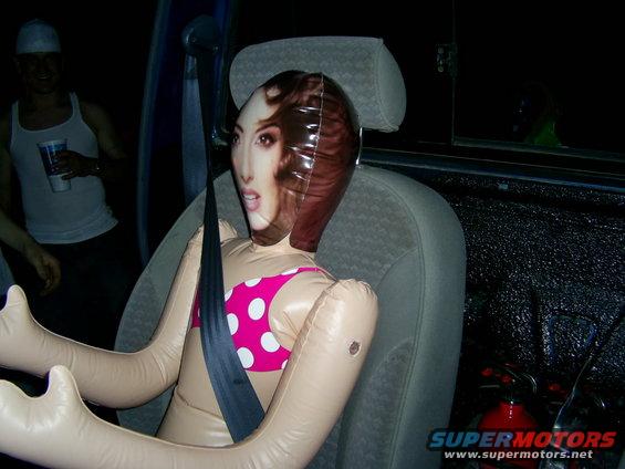 inflatable doll in car seat, wearing pink polka dot bikini, seatbelt on. guy in background holding cup