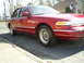 1995 Ford Crown Victoria - A red sedan is parked on the side of a residential street, with a house visible in the background.
