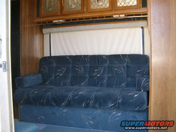 Interior of a trailer showing a blue sofa with a patterned fabric, wooden cabinets above, and a window with a pulled-down shade