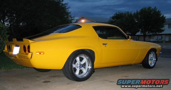 Yellow muscle car parked outside at dusk. It has a black stripe on the rear spoiler and chrome wheels