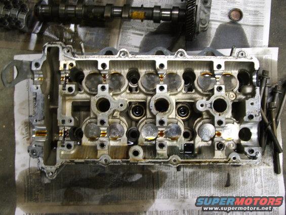 Disassembled engine cylinder head with camshaft and valves visible, resting on newspaper