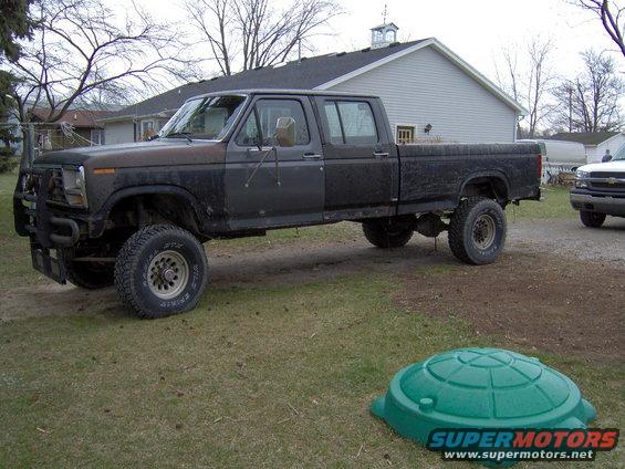 Dark gray crew cab pickup truck with a brush guard, lifted suspension, and dual rear wheels. The truck appears weathered