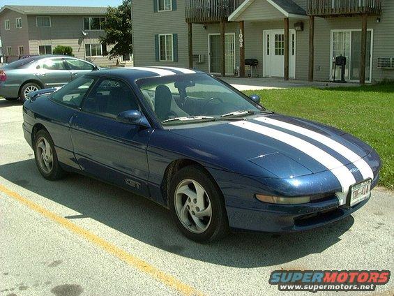 Blue 1995 Ford Probe with white racing stripes parked in front of an apartment building. Another car is parked behind it