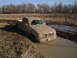 1981 Ford Bronco - A pickup truck is stuck in a muddy ditch, with its tires submerged in water and mud.