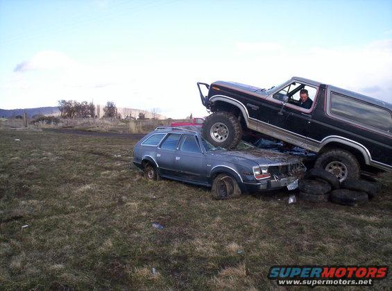 Black Ford Bronco on top of a crushed AMC station wagon in a grassy field