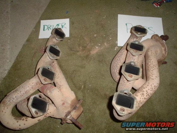 Two rusty exhaust manifolds, labeled 'DRIVER' and 'PASS', lie on a carpeted surface
