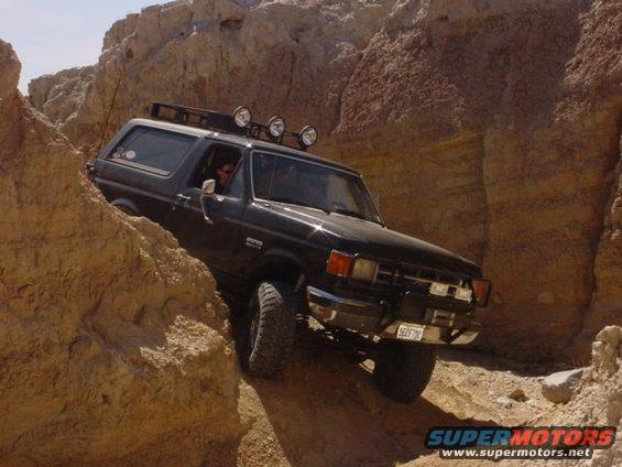 Black 1987 Ford Bronco navigating a rocky canyon, front-mounted lights on roof rack
