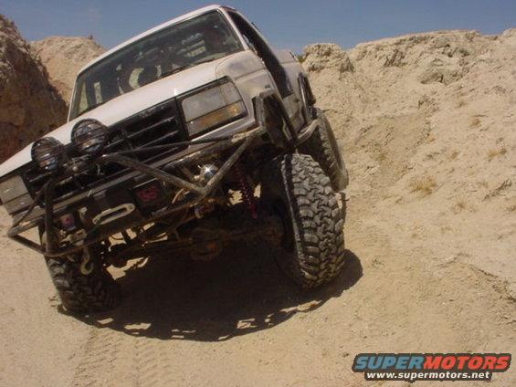 White 1988 Ford Bronco with off-road modifications driving on a dirt trail