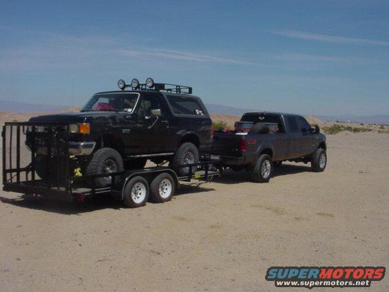 Black 1987 Ford Bronco on a trailer, front bull bar, roof rack with lights, towed by a dark gray pickup truck in a desert landscape