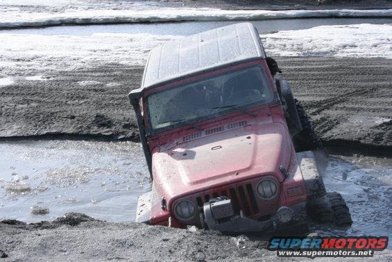 Red Jeep Wrangler stuck in muddy water, front end submerged. Winch visible on bumper