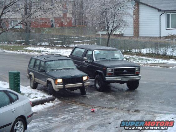 Black 1980s Ford Bronco parked on a wet street next to a Jeep Cherokee