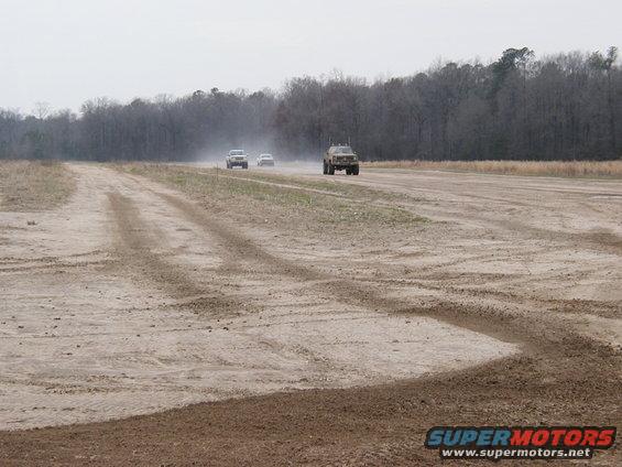 Three vehicles driving on a dusty dirt road with trees in the background