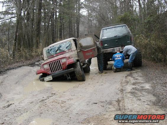 Red Jeep Wrangler stuck in mud, door open, with a green SUV behind it and people working on the SUV