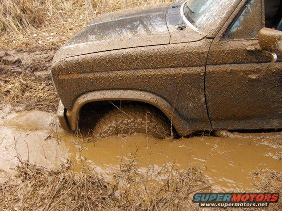 Mud-covered 1980s era truck or SUV stuck in a muddy puddle, front wheel submerged